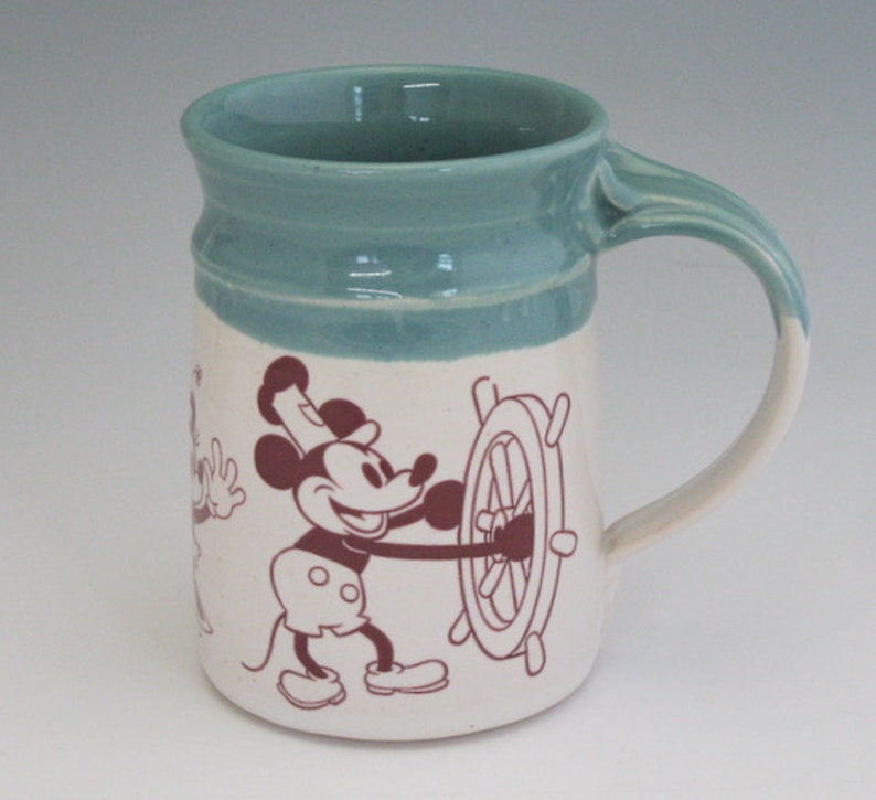 May include: A ceramic mug with a teal glaze and a white design featuring Mickey Mouse and a ship's wheel. Mickey is wearing a captain's hat and holding the wheel.