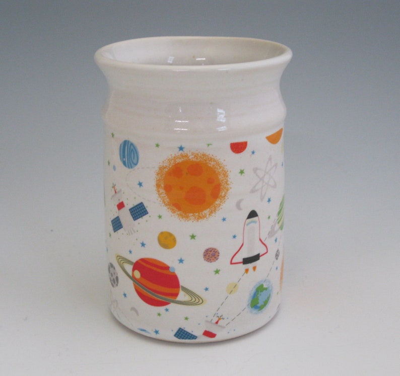 May include: White ceramic mug with a space-themed design. The mug features a pattern of planets, stars, rockets, and satellites in shades of orange, red, blue, and green.