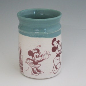 May include: A white ceramic mug with a light blue rim. The mug features a vintage illustration of Mickey Mouse and Minnie Mouse in black and red.