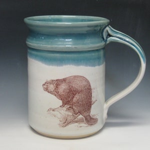 May include: A white ceramic mug with a teal glaze and a brown illustration of a beaver on the front. The mug has a rounded handle.