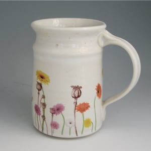 Puede incluir: Taza de cerámica blanca con un diseño floral con flores amarillas, naranjas, rosas y marrones. La taza tiene un asa y un borde ancho.