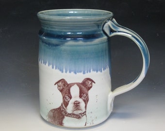 May include: A white ceramic mug with a blue and green glaze. The mug has a handle and features a brown and white illustration of a Boston Terrier dog.