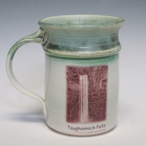 May include: A white ceramic mug with a green glaze and a picture of Taughannock Falls in a brown tone. The mug has a handle and the text "Taughannock Falls" is printed on the bottom.