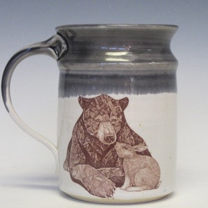 May include: A ceramic mug with a gray glaze and a brown illustration of a bear and a rabbit. The bear is lying down and the rabbit is sitting next to it.