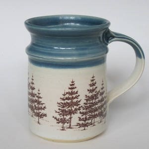 May include: A white ceramic mug with a blue rim and brown tree silhouettes. The mug has a large handle.