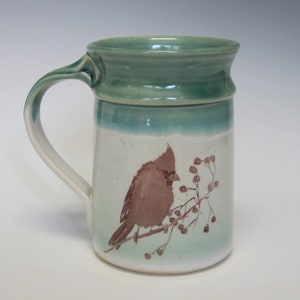 May include: A green ceramic mug with a brown cardinal bird illustration and a branch of berries. The mug has a white background with a green glaze on the top and bottom.