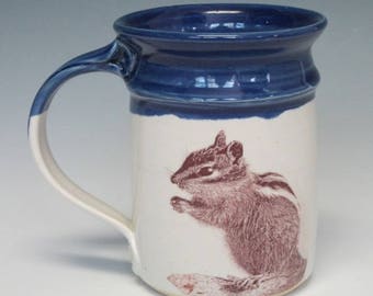 May include: A ceramic mug with a white body and a dark blue glazed rim and handle. The front of the mug features a detailed brown illustration of a chipmunk in a seated position. The mug has a cylindrical shape with a flared top edge.