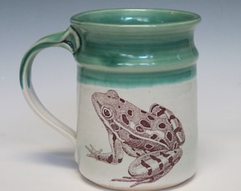 May include: A white ceramic mug with a green glaze around the top. The mug has a brown illustration of a frog on the front.