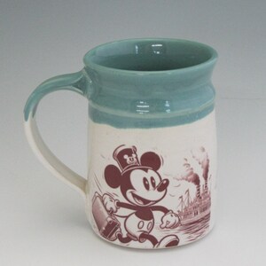 May include: A white ceramic mug with a blue rim and a vintage illustration of Mickey Mouse carrying a suitcase. The illustration is in brown and shows Mickey Mouse walking towards a steamboat in the background.