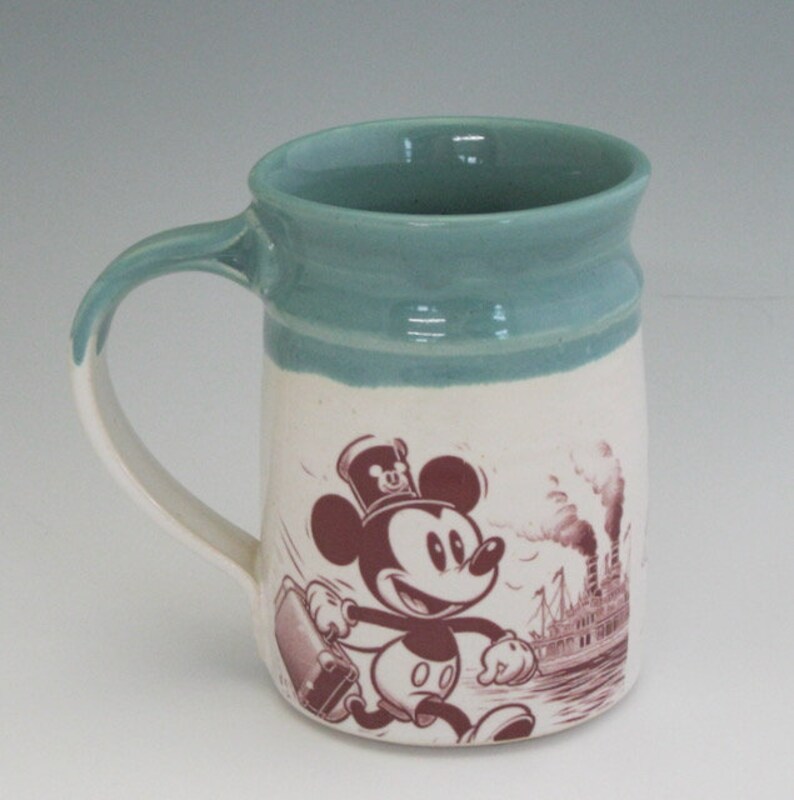 May include: A white ceramic mug with a blue rim and a vintage illustration of Mickey Mouse carrying a suitcase. The illustration is in brown and shows Mickey Mouse walking towards a steamboat in the background.