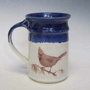May include: A ceramic mug with a blue rim and handle. The mug features a painted image of a brown cardinal bird perched on a branch. The background is white. The mug is designed for hot beverages.