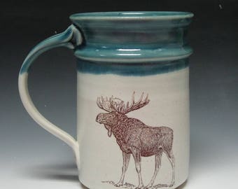 May include: A white ceramic mug with a teal blue rim and a brown moose illustration. The mug has a large handle.