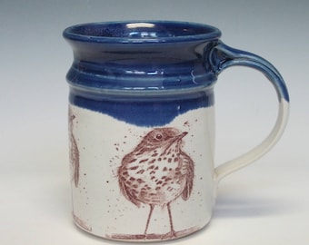 May include: A white ceramic mug with a blue rim and handle. The mug features a brown and white illustration of a bird with a speckled breast.