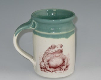 May include: A ceramic mug with a green glaze and a white band around the middle. The mug features a detailed illustration of a brown toad sitting on a white background.