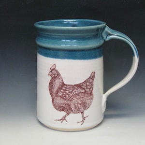 May include: A white ceramic mug with a teal blue rim and handle. The mug has a brown line around the top and a brown illustration of a chicken on the front.