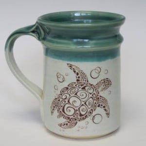 May include: A white ceramic mug with a green rim and a brown sea turtle design. The turtle is surrounded by bubbles.