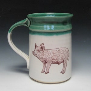 Könnte beinhalten: Eine weiße Keramiktasse mit grünem Rand und einer braunen Illustration eines Schweins auf der Vorderseite.