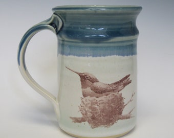 May include: A ceramic mug with a blue glaze and a brown hummingbird illustration. The hummingbird is perched on a branch with a nest.