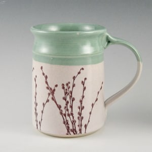 May include: A ceramic mug with a light green glaze and a white band with a brown floral design. The mug has a rounded handle.