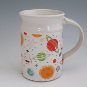 May include: White ceramic mug with a space-themed design. The mug features planets, stars, rockets, and an atom symbol in shades of red, orange, yellow, green, blue, and gray.