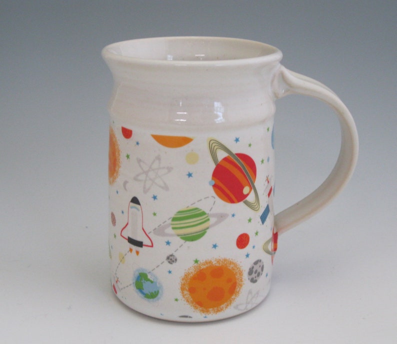 May include: White ceramic mug with a space-themed design. The mug features planets, stars, rockets, and an atom symbol in shades of red, orange, yellow, green, blue, and gray.