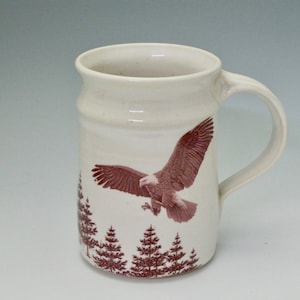 May include: White ceramic mug featuring a brown image of a bald eagle in flight over a forest. The eagle's wings are spread wide, and the forest is composed of pine trees.