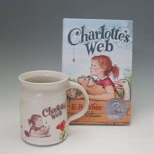 May include: A cream-colored ceramic mug and a hardcover book titled "Charlotte's Web." The mug features a graphic of a girl and a pig, with the book's title. The book cover shows a girl, pig, and goose.
