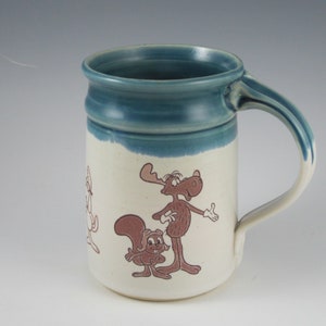 May include: A white ceramic mug with a blue glaze around the rim. The mug features a cartoon illustration of a moose and a squirrel. The moose is brown and has a long snout and large ears. The squirrel is brown and has a bushy tail. The mug has a handle on the right side.