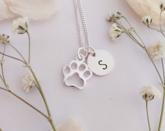 May include: Silver necklace with a paw print charm and a small disc charm with the letter 'S' engraved on it. The necklace is displayed on a white background with white flowers.