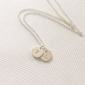 Puede incluir: Un collar de cadena de plata con dos pequeños discos de plata, uno con la letra "T" y el otro con la letra "K" colgando de él.