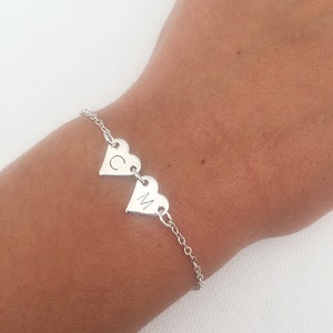 May include: A silver bracelet featuring two heart-shaped charms. Each charm has a single initial engraved on it. The bracelet is worn on a wrist, and the chain is delicate and links the charms together.