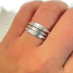 Könnte beinhalten: Ein silberner Ring mit drei Bändern, die jeweils mit einem Namen und einem Datum graviert sind. Das obere Band trägt die Inschrift "Alfie 18.01.2023", das mittlere Band die Inschrift "Melissa" und das untere Band ist leer.