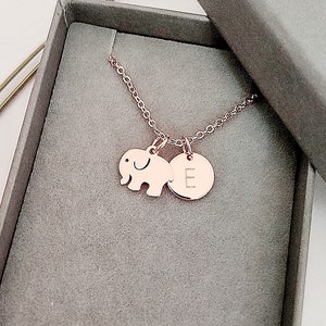 Könnte beinhalten: Eine Halskette aus Roségold mit einem kleinen Elefanten-Anhänger und einem runden Anhänger mit dem eingravierten Buchstaben "E". Die Halskette befindet sich in einer grauen Geschenkbox.