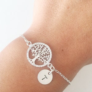 Puede incluir: Una pulsera de plata con un dije de árbol de la vida y un dije de letra 'T'. El dije de árbol de la vida está rodeado de pequeñas piedras transparentes.