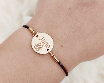May include: A black cord bracelet with a gold round charm that says "Juno" with a paw print and a heart inside the print.