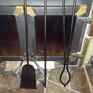 May include: A set of four black fireplace tools hanging on a wall. The tools include a poker, tongs, a shovel, and a brush. The tools are made of metal and have a simple design.