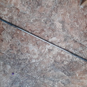 May include: A long, silver metal hook with a loop at one end and a pointed end at the other. The hook is lying on a tiled floor.