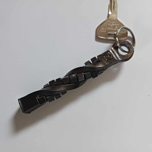 May include: A black metal keychain with a twisted design and a square pattern. The keychain is attached to a silver key with a key ring.