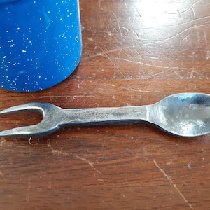 May include: A metal spoon with a fork on the end. The spoon is silver and the fork is black.