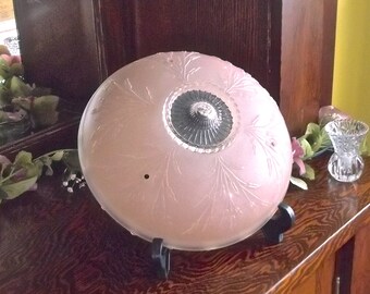 May include: A pink glass ceiling light fixture with a decorative etched design. The light fixture is on a wooden surface with a floral arrangement.