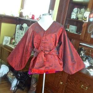 K&ouml;nnte beinhalten: Ein rotes und violettes Kimono mit Blumenmuster. Das Kimono hat lange &Auml;rmel und eine Sch&auml;rpe, die sich in der Taille bindet. Das Kimono wird auf einer Schaufensterpuppe pr&auml;sentiert.
