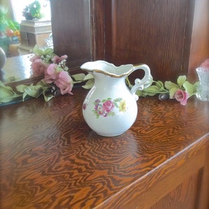 Peut inclure: Un pichet en porcelaine blanche avec un motif floral et une bordure dor&eacute;e. Le pichet a une anse et un bec verseur. Il est pos&eacute; sur une surface en bois.