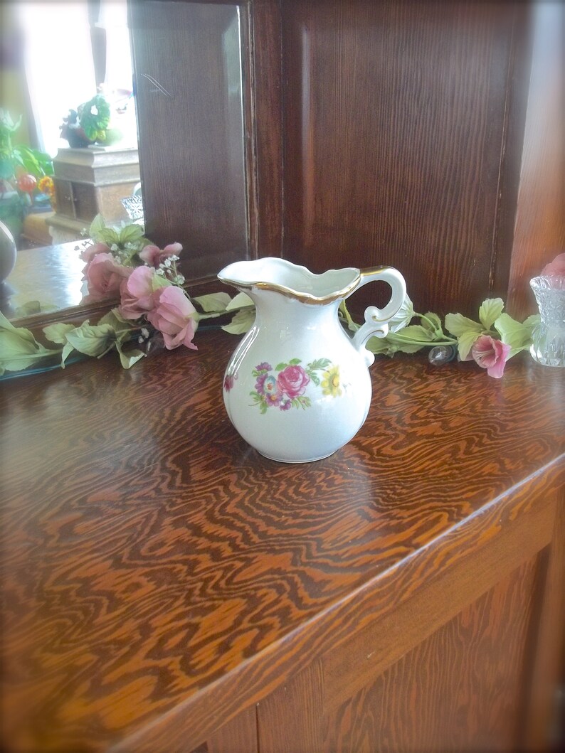 Peut inclure: Un pichet en porcelaine blanche avec un motif floral et une bordure dor&eacute;e. Le pichet a une anse et un bec verseur. Il est pos&eacute; sur une surface en bois.