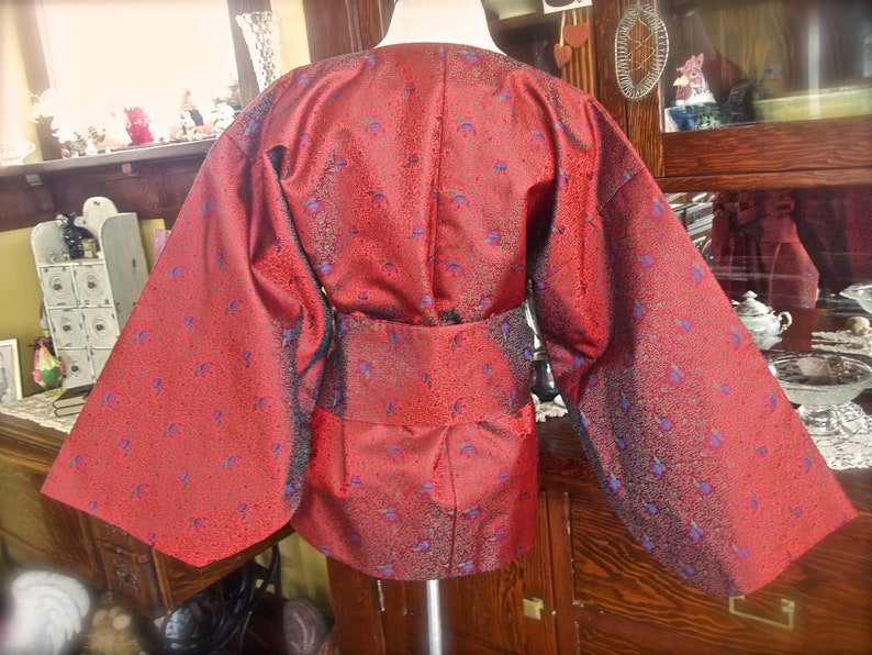 K&ouml;nnte beinhalten: Ein rotes Kimono mit einem blauen und goldenen Blumenmuster. Das Kimono hat lange &Auml;rmel und einen G&uuml;rtel.