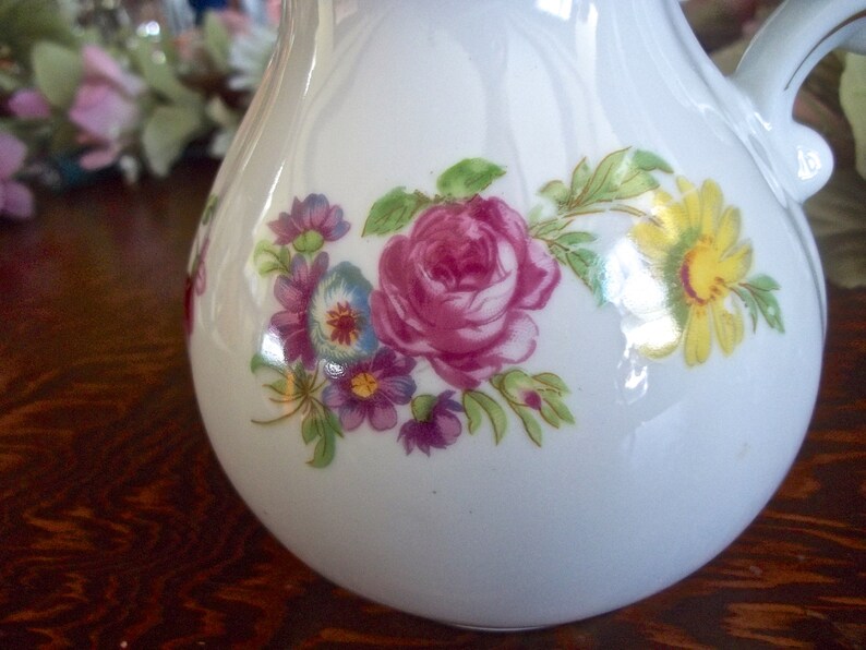 Peut inclure: Pichet en c&eacute;ramique blanche avec un motif floral compos&eacute; de fleurs roses, jaunes et violettes. Le pichet a une anse et un corps en forme de poire.