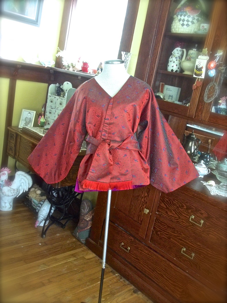 K&ouml;nnte beinhalten: Ein rotes und violettes Kimono mit Blumenmuster. Das Kimono hat lange &Auml;rmel und eine Sch&auml;rpe, die sich in der Taille bindet. Das Kimono wird auf einer Schaufensterpuppe pr&auml;sentiert.