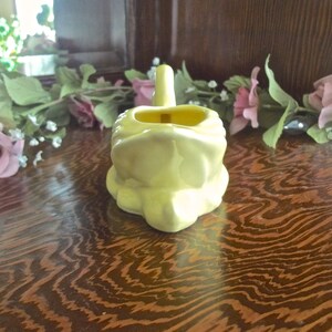 May include: A pale yellow ceramic frog-shaped container with a handle. The container has a rectangular opening and sits on a wooden surface. Artificial pink roses and greenery are in the background.