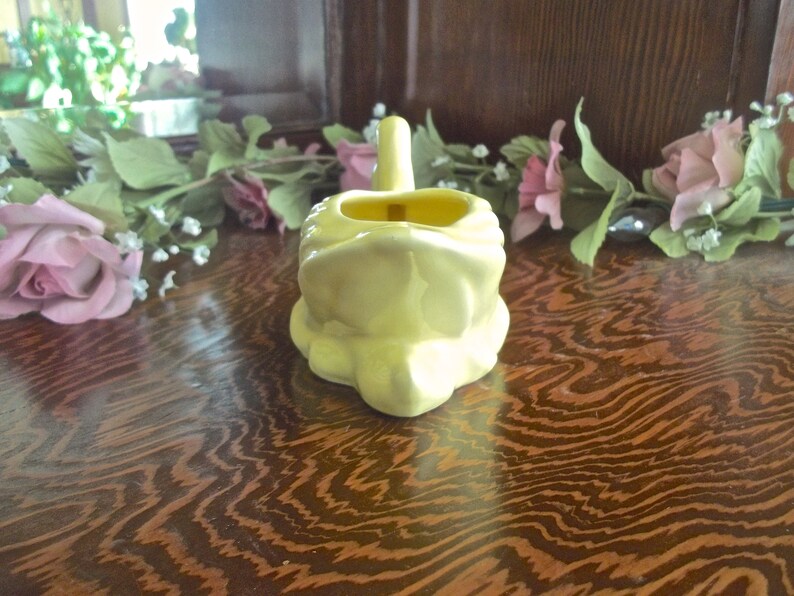 May include: A pale yellow ceramic frog-shaped container with a handle. The container has a rectangular opening and sits on a wooden surface. Artificial pink roses and greenery are in the background.