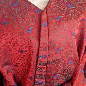 K&ouml;nnte beinhalten: Rote Seidenrobe mit einem detaillierten Muster aus gr&uuml;nen und blauen Symbolen. Die Robe hat einen V-Ausschnitt und einen Knopfverschluss.