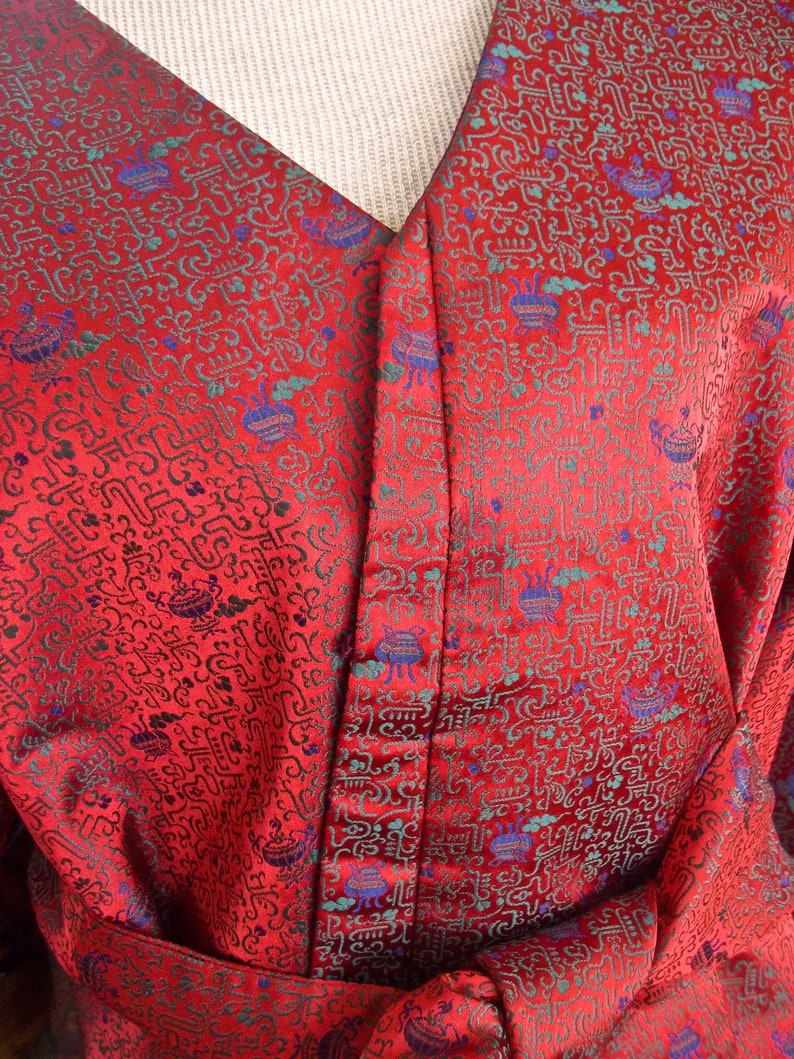 K&ouml;nnte beinhalten: Rote Seidenrobe mit einem detaillierten Muster aus gr&uuml;nen und blauen Symbolen. Die Robe hat einen V-Ausschnitt und einen Knopfverschluss.